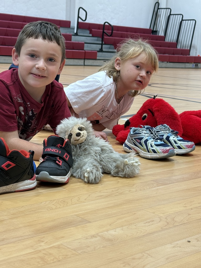 Two kids sitting on a basketball court, one wearing sneakers, the other lying with stuffed animals.
