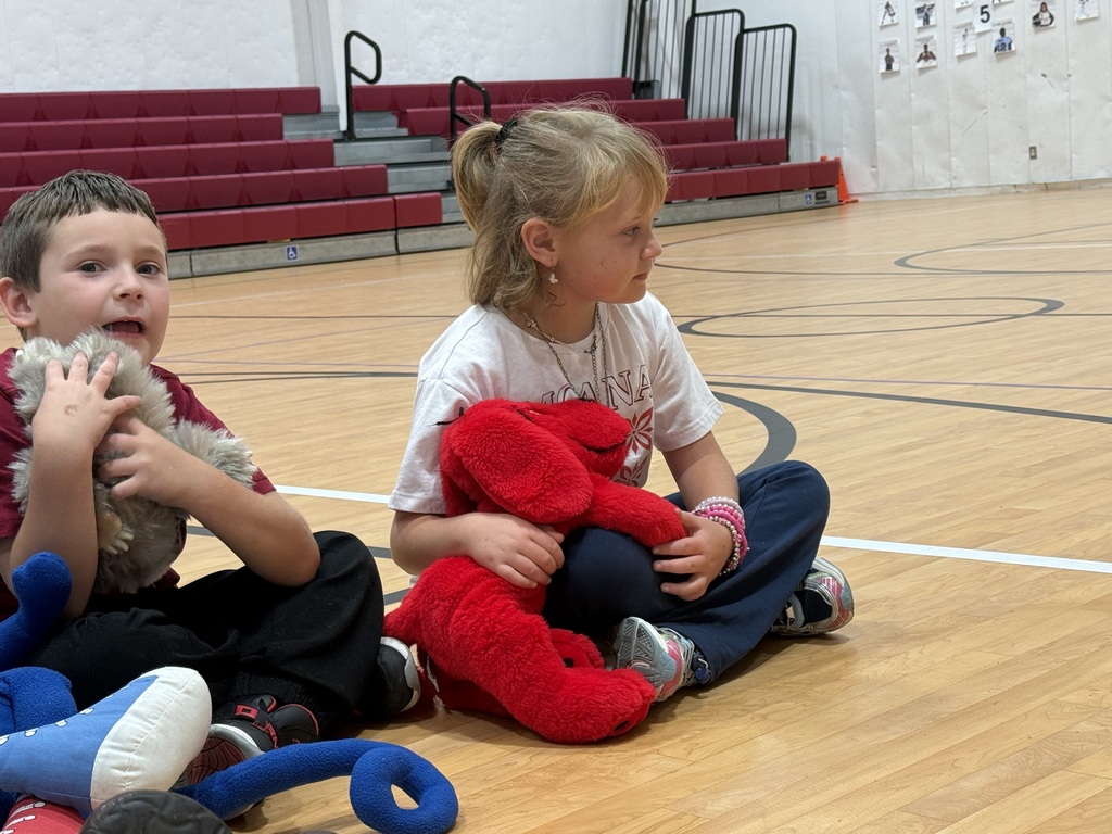Children sit on a basketball court, hugging stuffed animals, with red bleachers in the background.
