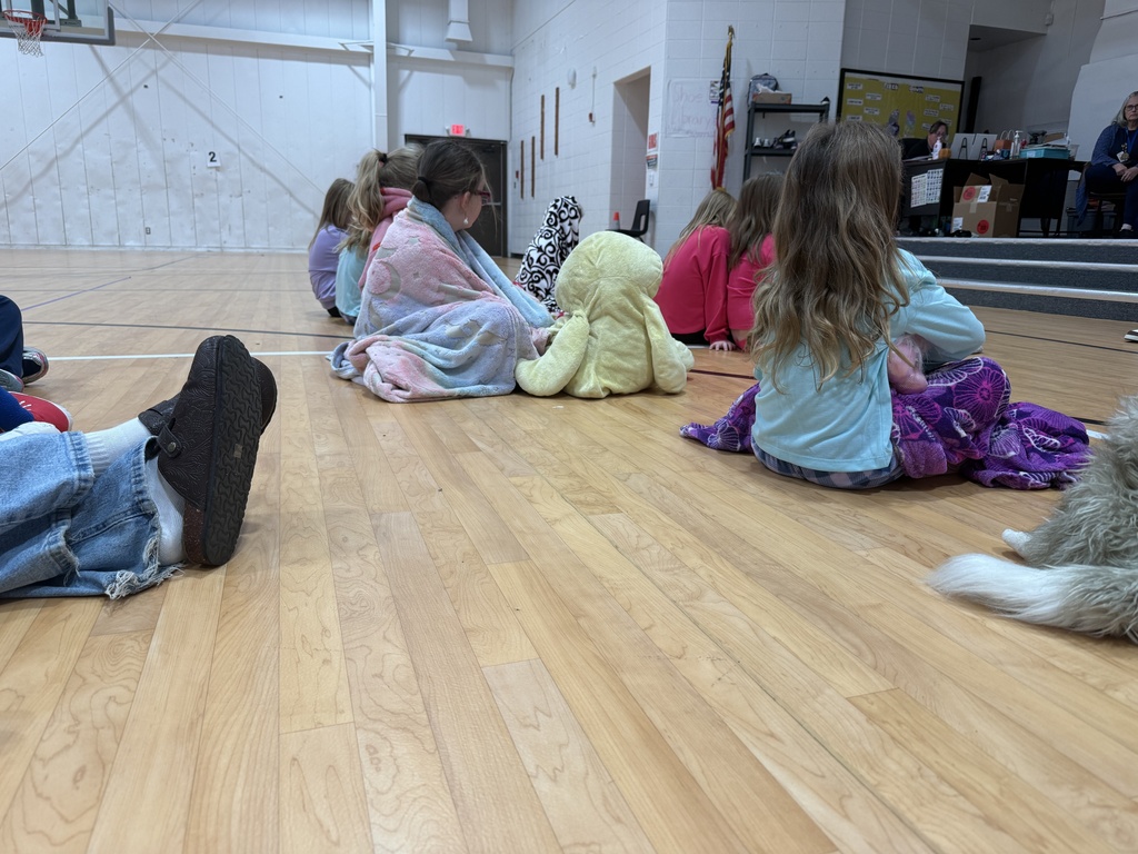 Children sitting in a circle on a gym floor with some wearing blankets and a stuffed toy