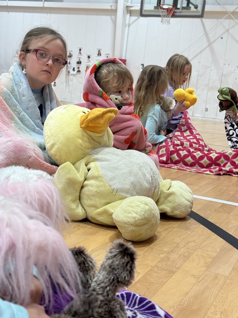 A group of children sitting in a gymnasium, holding stuffed animals. Some children have blankets around them.