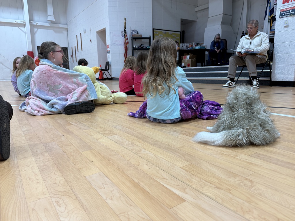 Several children sit on a floor, wrapped in blankets. One person sits in a chair reading a book.