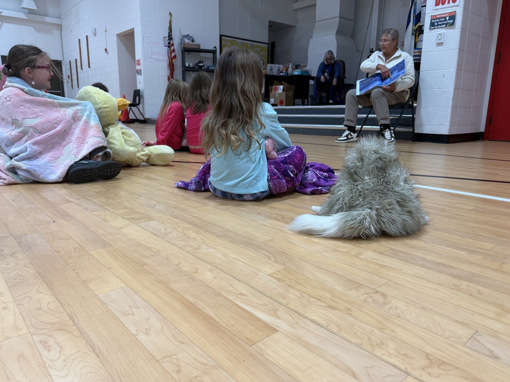 Children sit on the floor with stuffed animals, one holding a book. A person sits in a chair on a stage.