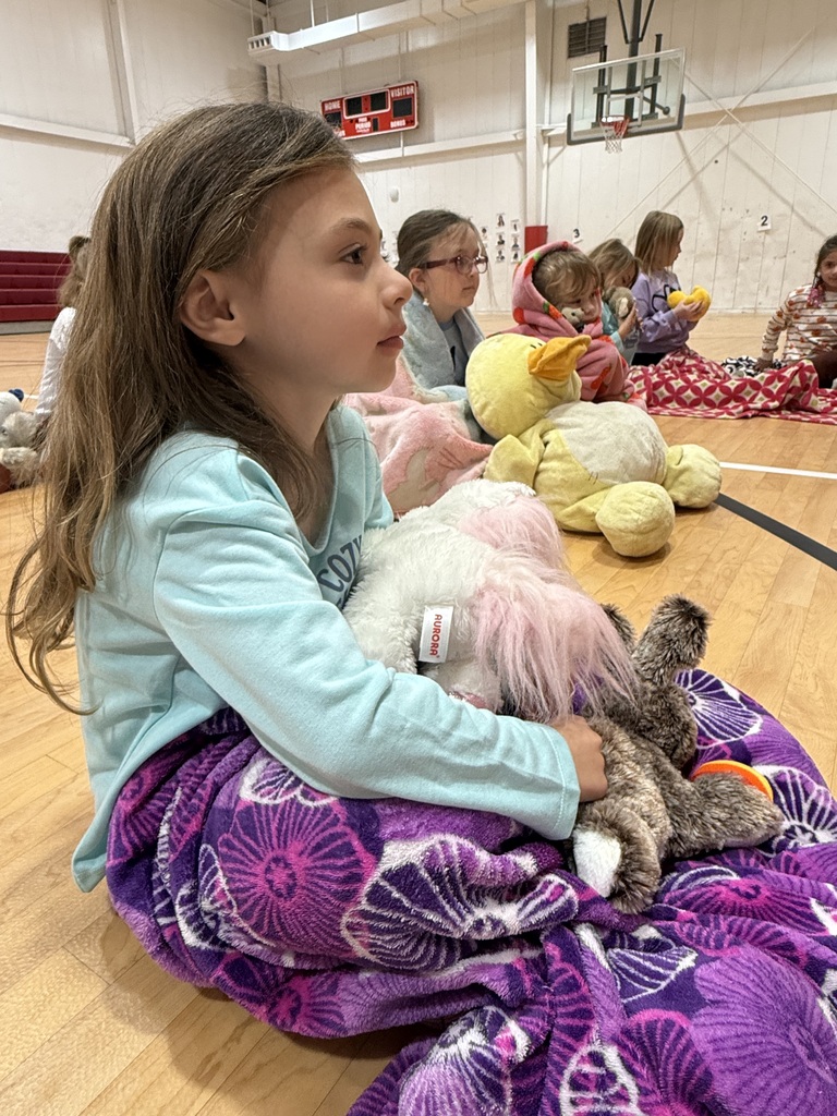A young girl sits on a gymnasium floor, holding a stuffed animal while others sit around her.