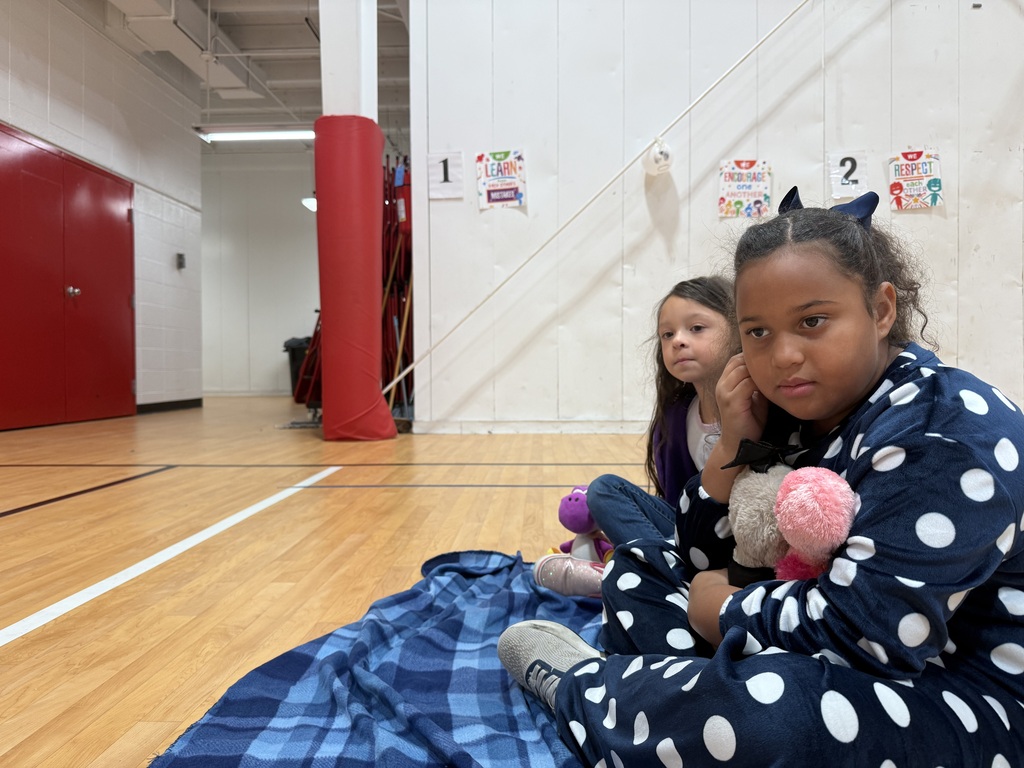 Two young girls sit on a blanket on a gym floor, one holding a stuffed animal.