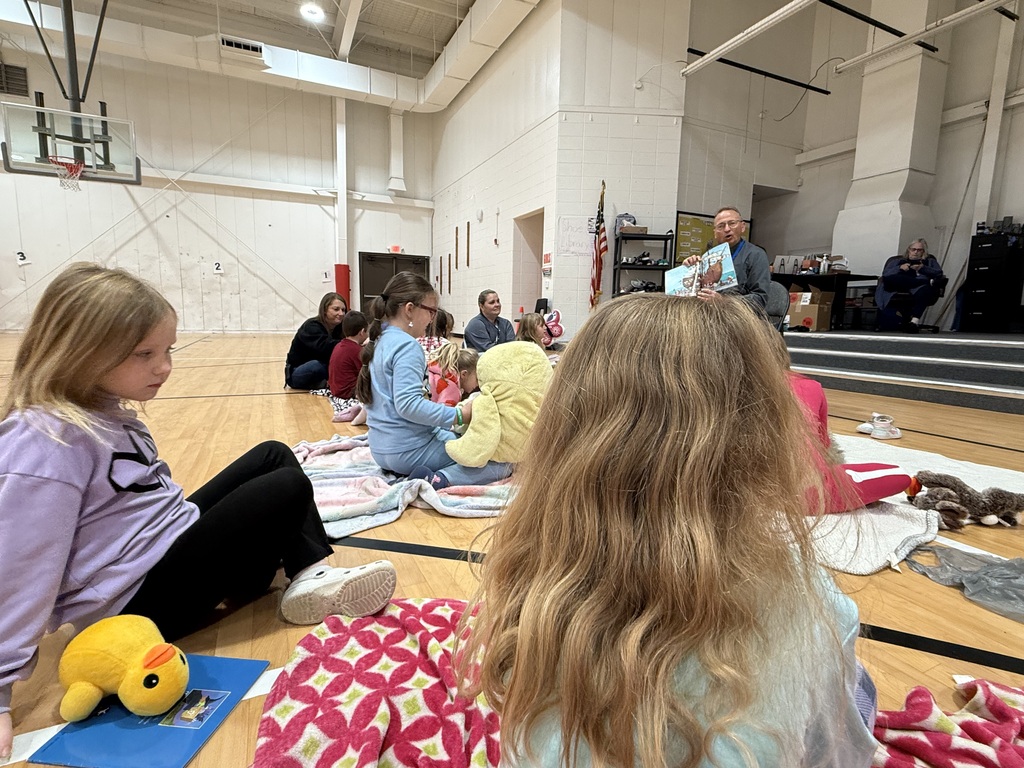 Several students seated on blankets on a gym floor, with a person holding a yellow stuffed animal.