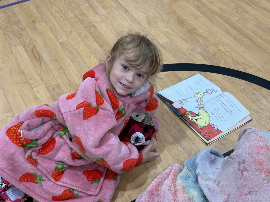 Child in pink pajamas with strawberry pattern sitting on floor, reading book and holding stuffed toy.