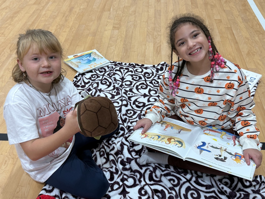 Two girls sit on a patterned rug, one holding a book and the other holding a turtle toy.