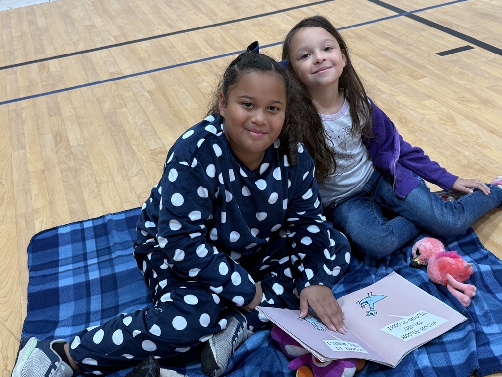 Two girls sit on a blanket, one reading a book, the other smiling, in a gym.