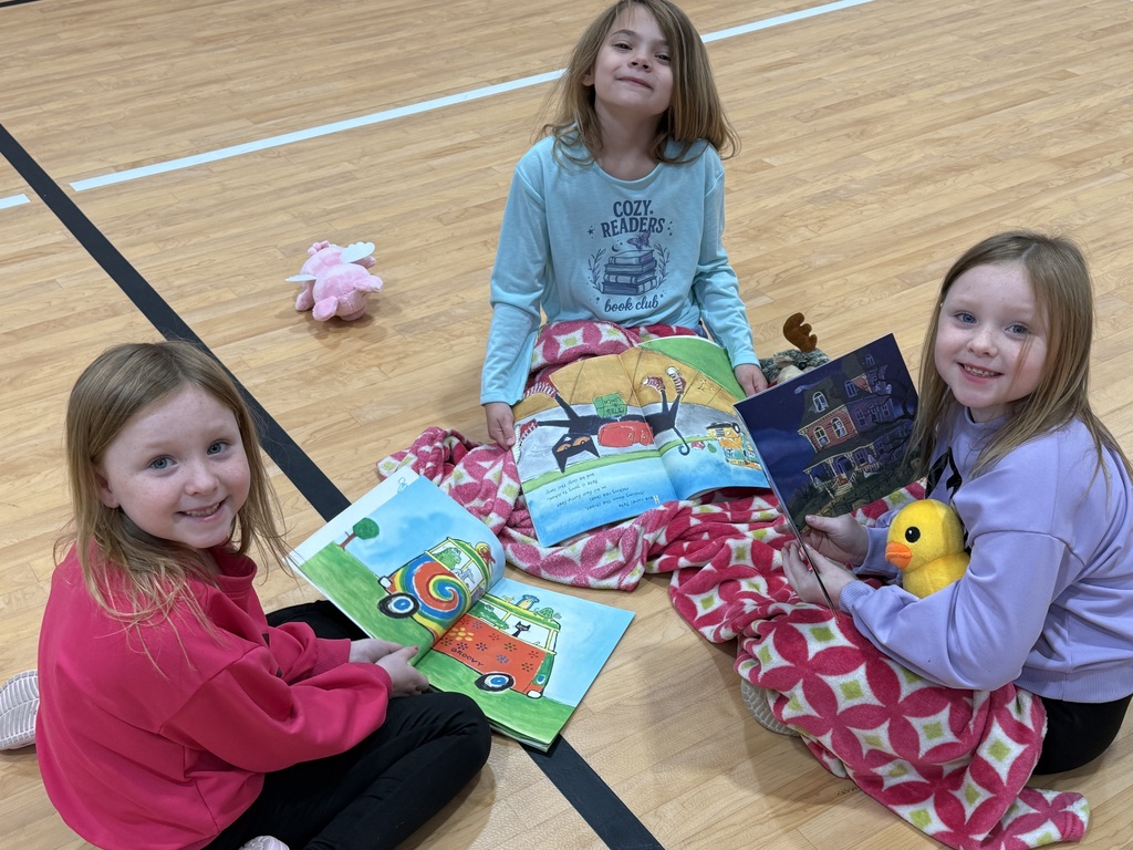 Three young girls sit on a gym floor, reading books and holding toys, with a gym floor as background.