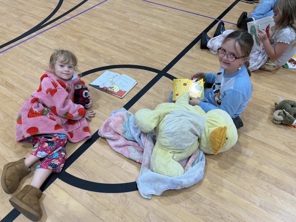 Two children sit on the floor in a gym, reading books and holding stuffed animals.