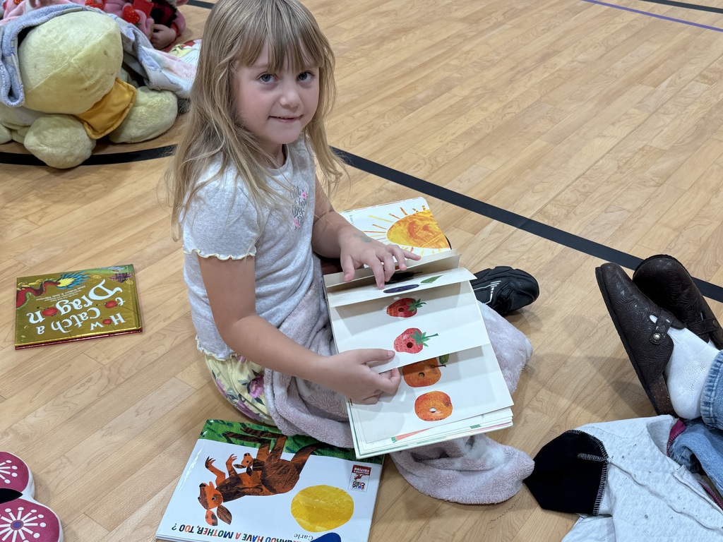 Girl holding an open book while sitting on a wooden floor in a gymnasium.