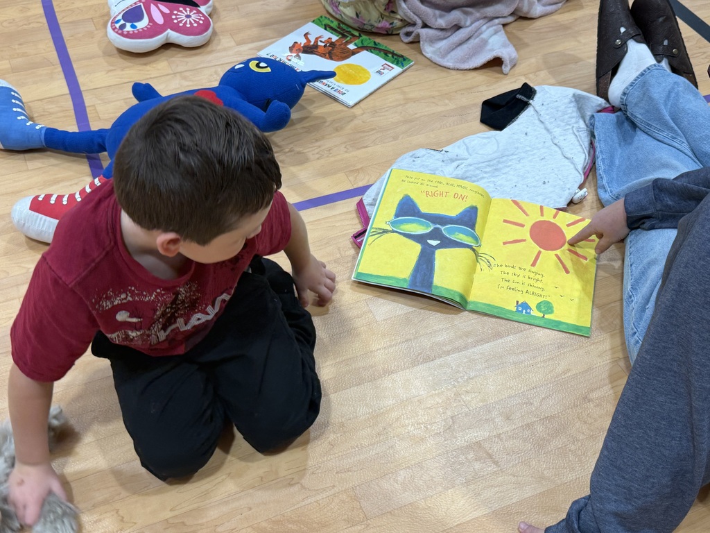 Child kneeling on a floor with an open book, pointing at images while another person points nearby.