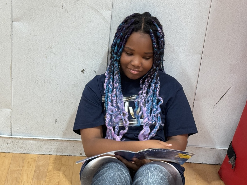 A young person with blue braids sitting on a floor, holding and reading a book.