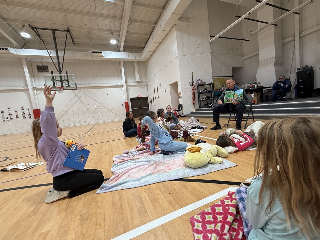 Children sit on blankets in a gymnasium. A woman reads a book to them. One woman holds a book.