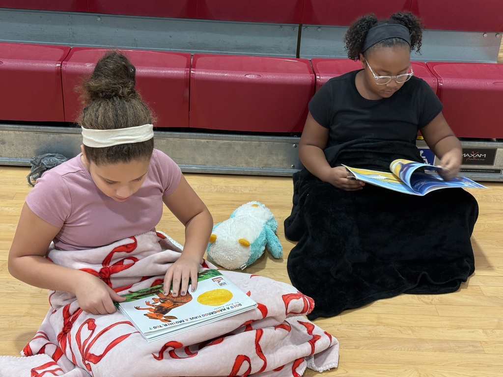 Two students sitting on a floor with books, one wearing a headband, other wearing glasses, reading.
