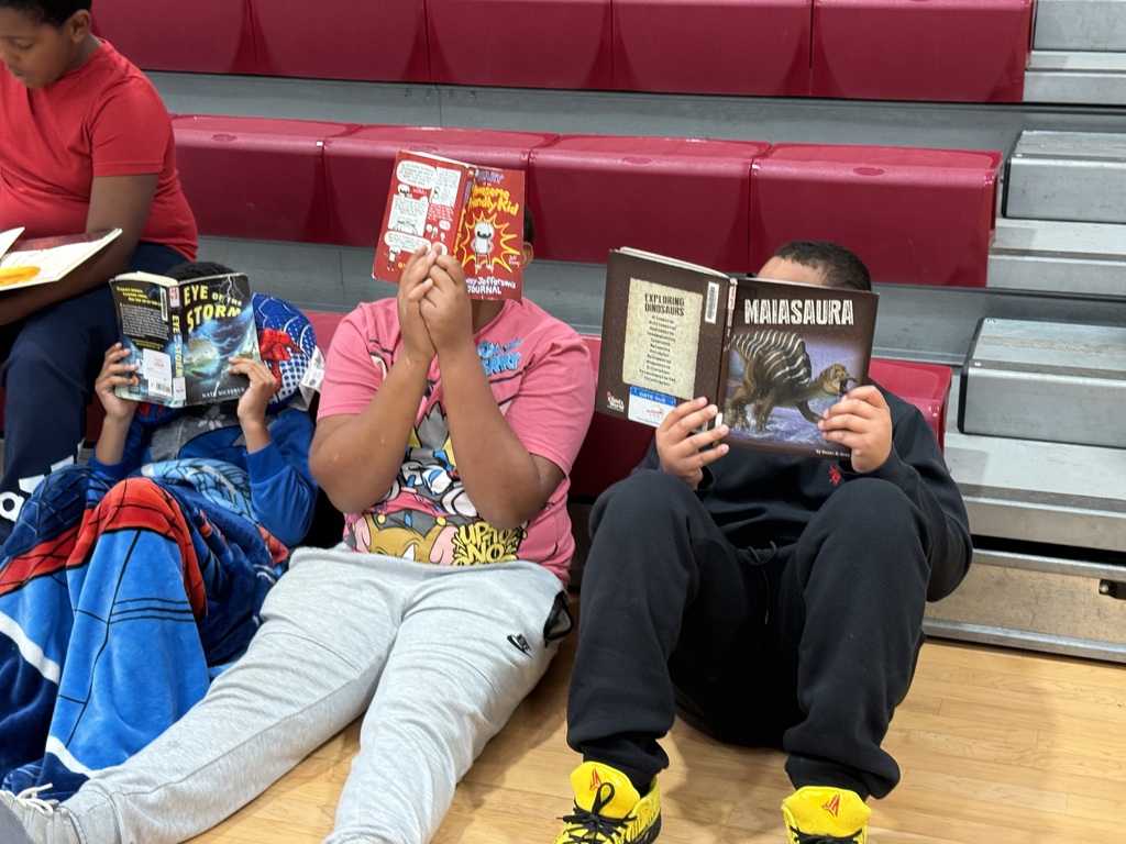 Three students read books on a gym floor with red bleachers in the background.