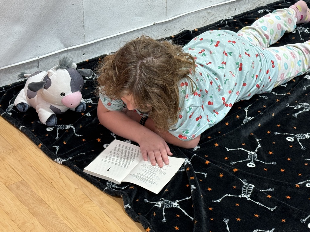 A young girl in pajamas lies on a black blanket with skeletons, reading a book.
