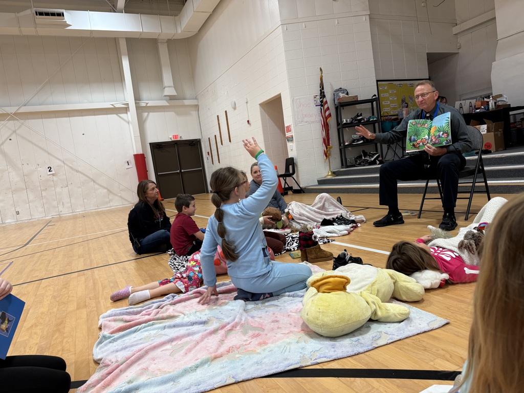 A man reads a book to young children in a gym while they sit on a blanket.