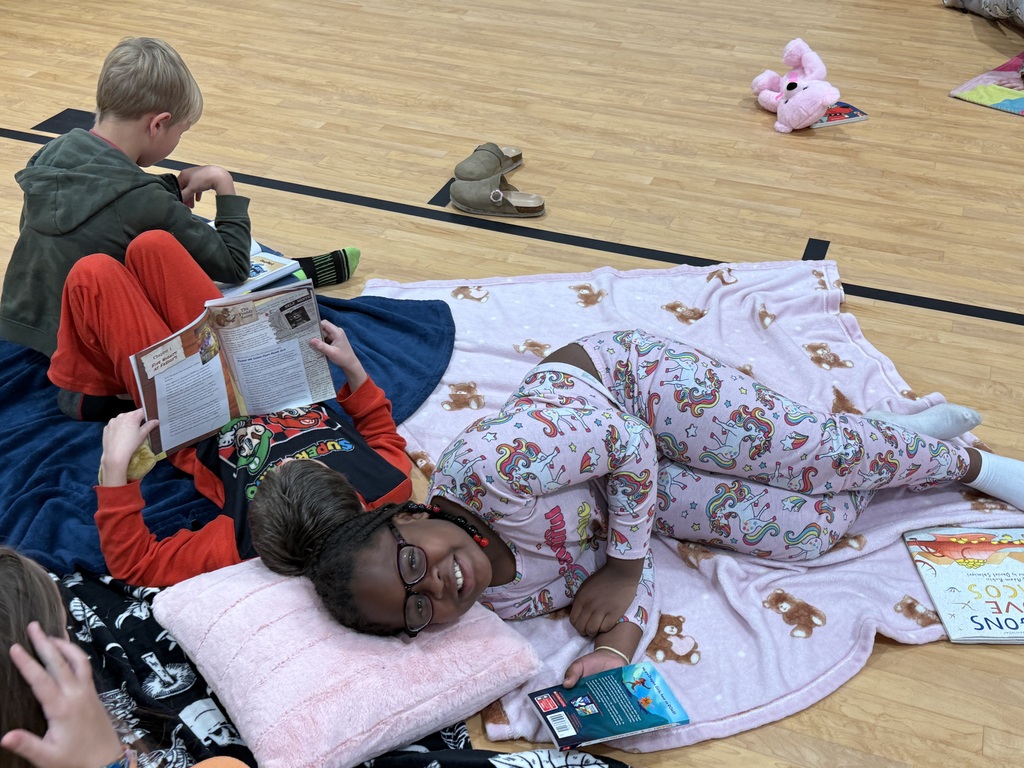 Students sitting on a gym floor with blankets, reading books and smiling; one child wearing glasses.