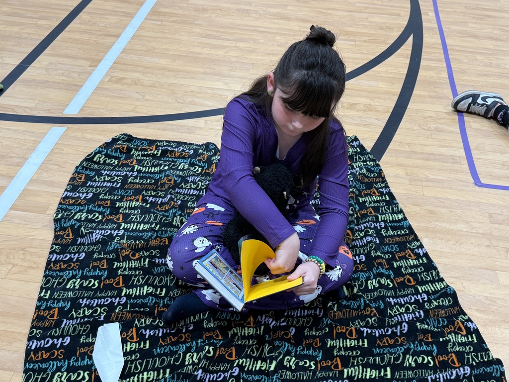 A student sits on a rug, holding a book and yellow folder, with sports court lines and a white line in the background.
