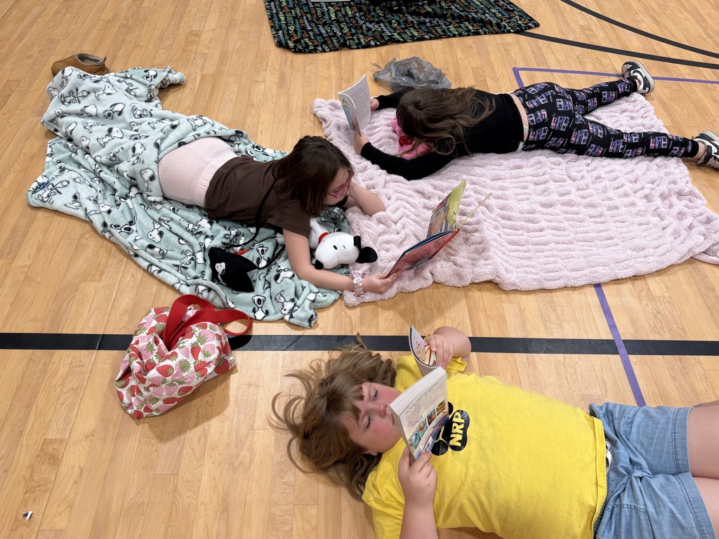 Three children lie on blankets on a gym floor, reading books, with a bag and stuffed toys nearby.