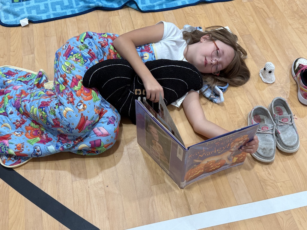 Child lying on floor reading book with shoes and blanket nearby. Wooden floor with black line.