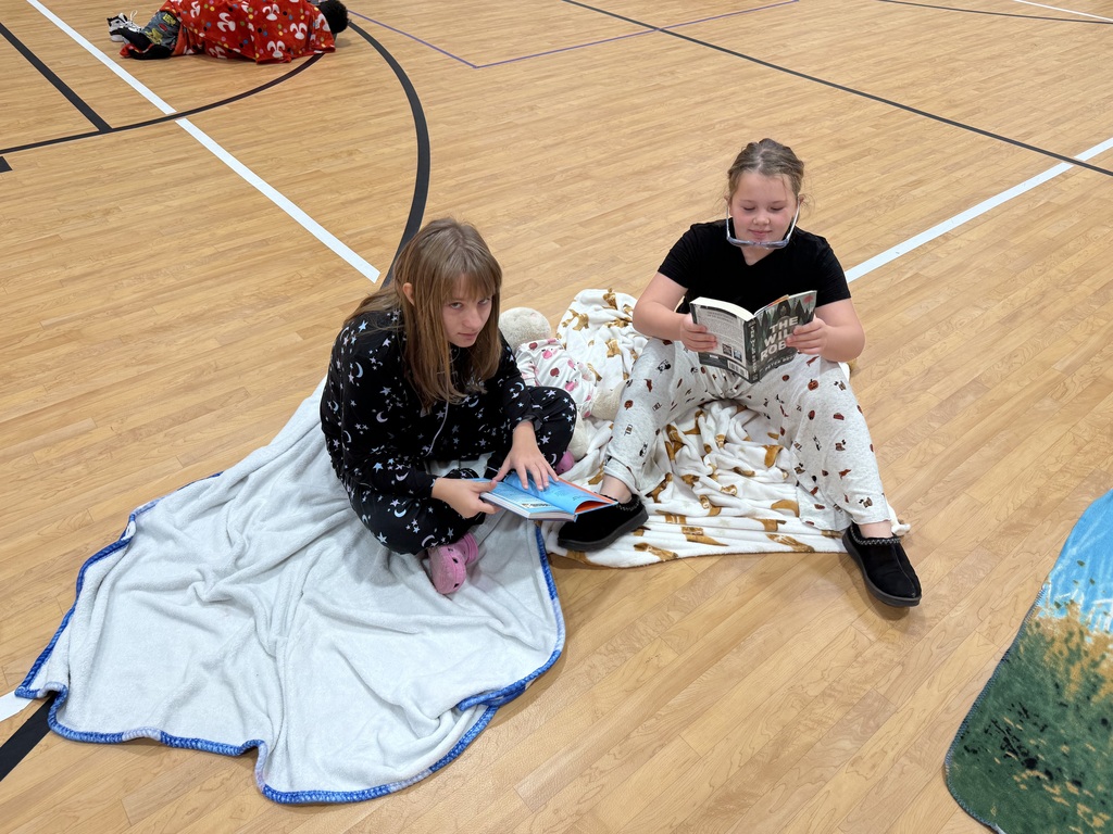 Two students sit on blankets in a gym. One reads a book while the other studies.