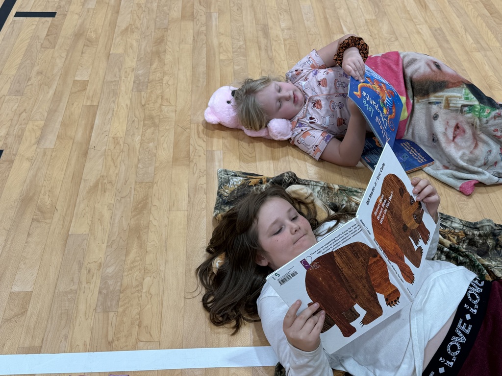 Two girls lie on floor, reading books, one holding a stuffed animal, on a wooden floor.