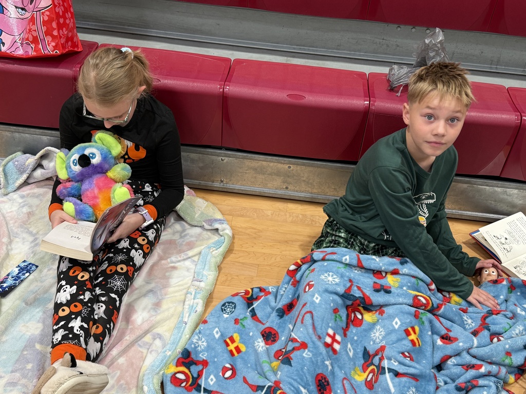 Two children reading books on a blanket on a gym floor with red seating in the background.