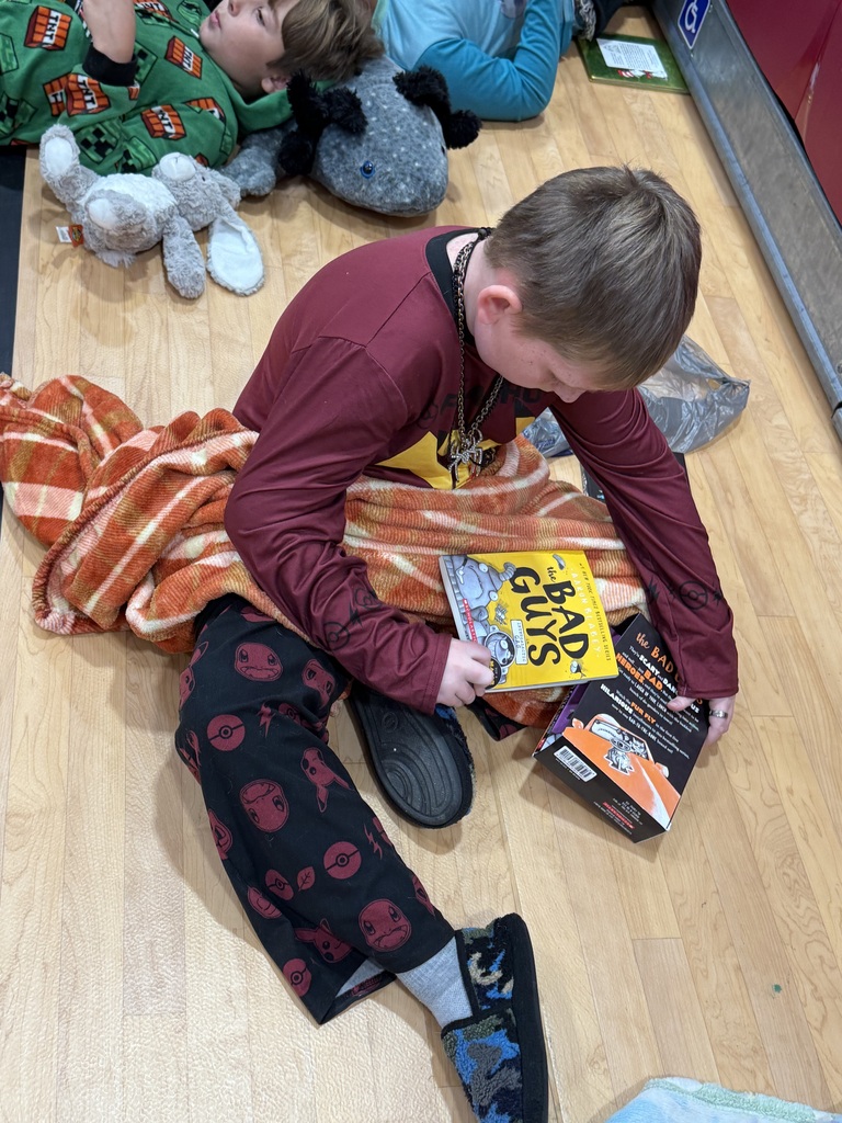 Boy reading a book while sitting on a wooden floor with stuffed animals. Another boy sleeps next to him.