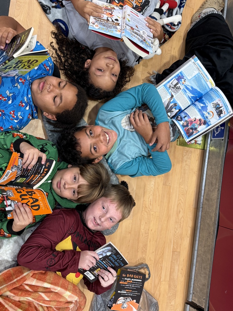 Five children lying on a wooden floor, holding books and looking at pictures; one is wearing pajamas.