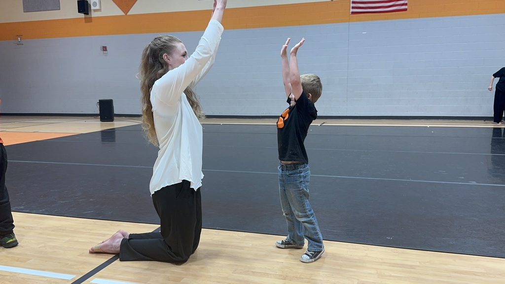 Two people practice stretching on a gym floor, one kneeling, one standing, with a basketball court visible.