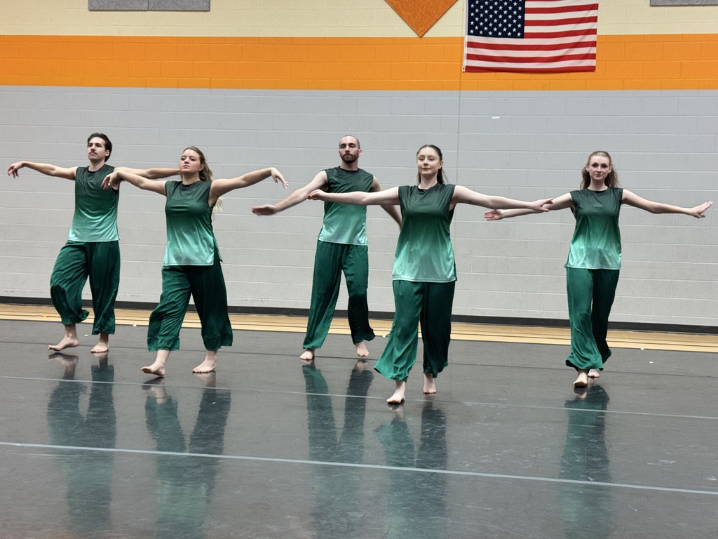 Five dancers in green outfits, barefoot, performing a routine on a shiny floor, with an American flag in background.