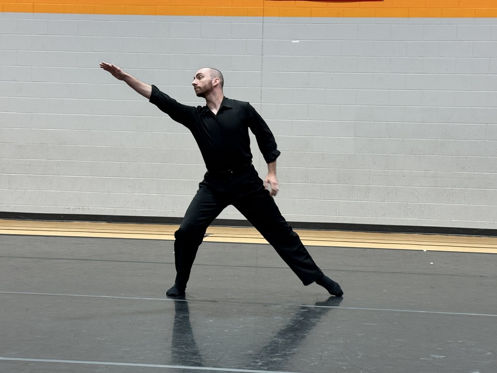 A man in a black outfit performs a dance move in a gym with gray walls.