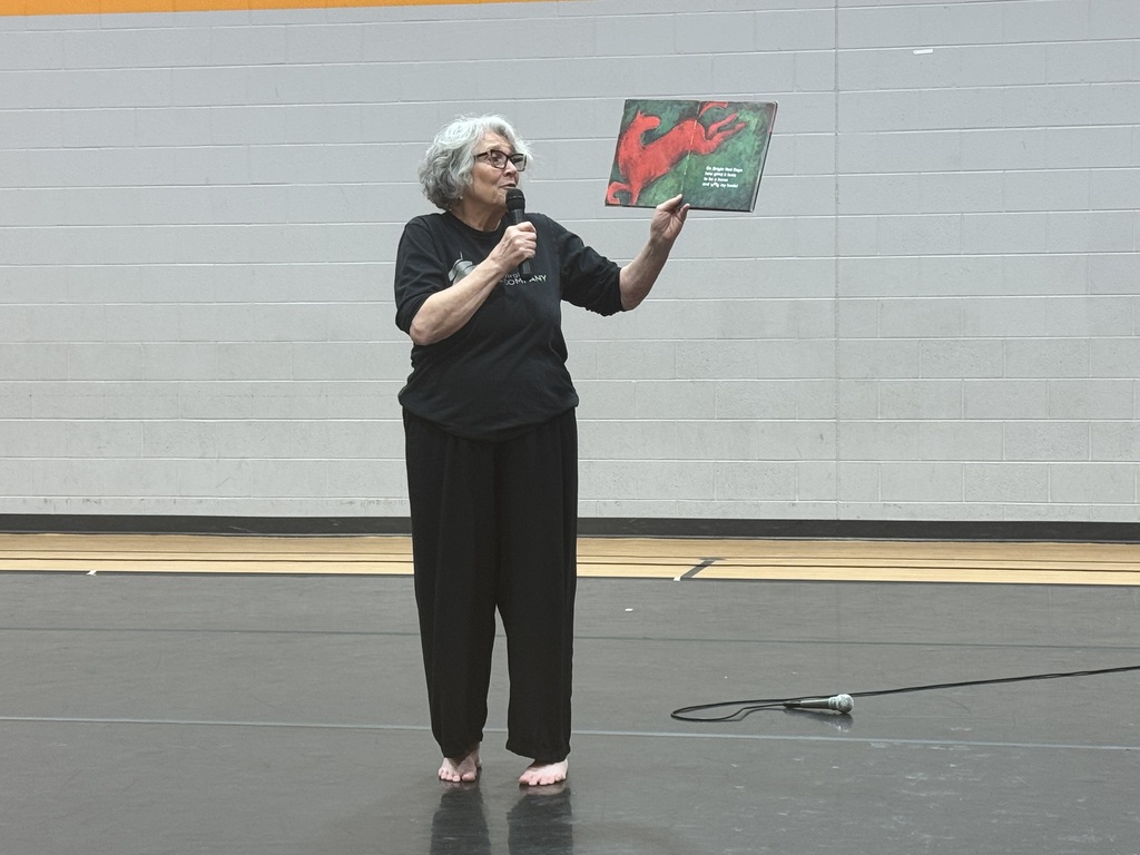 Woman with white hair speaks into microphone, holding book with red cover and black text, standing on stage.