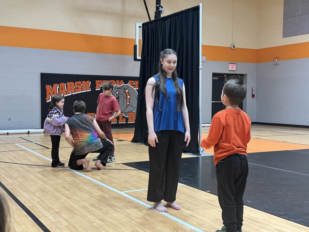 Children in a gym, one standing tall, others kneeling, one with their hands raised.