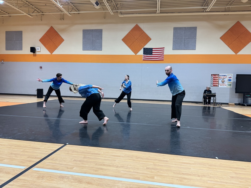 Four people in blue shirts and black pants on a gym floor, performing a routine.