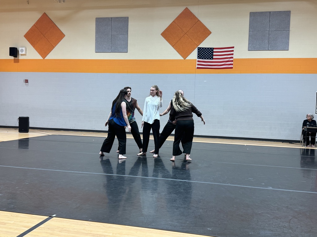 Four people in white tops and black pants dance in a gym. A U.S. flag hangs on the wall.