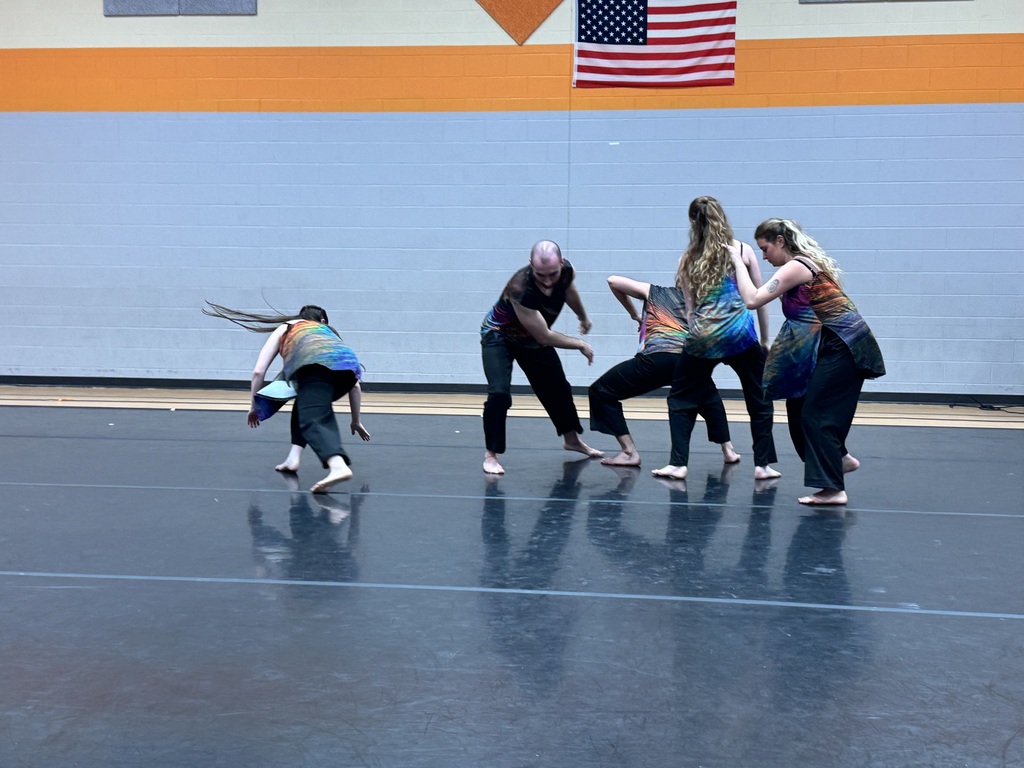 Several dancers performing a routine on a gym floor with a flag on the wall.