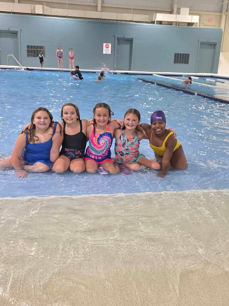 Six girls in swimsuits pose for a photo in a pool. The water is blue.
