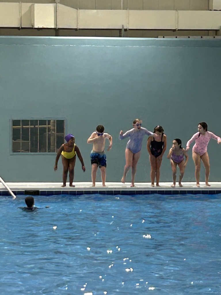 Eight swimmers in various swimwear colors stand on a pool deck, facing the pool. One swimmer swims in the pool.