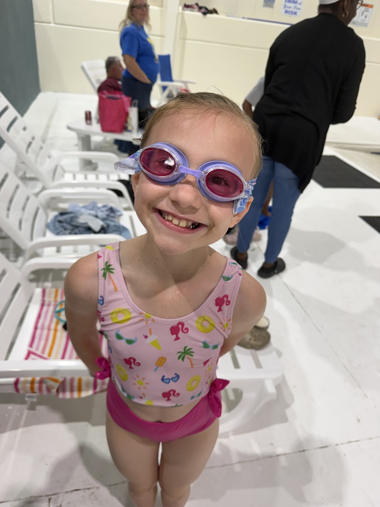 Girl wearing swimming goggles smiles inside a pool area, with chairs and other people in background.
