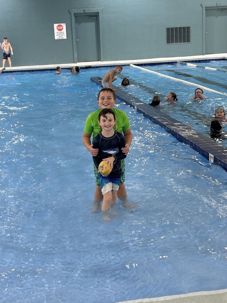 Two children standing in a pool, one holding a yellow ball. Other children swim in the background.