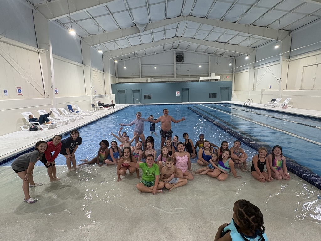 Group of children and adults sitting and standing in shallow end of indoor swimming pool with lane lines.