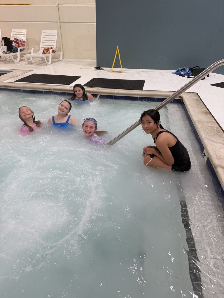 Five girls in swimsuits sitting in a swimming pool with water splashing. A woman in black swimsuit crouches by the pool edge.
