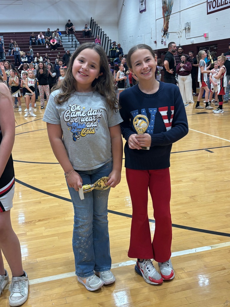 Two young girls on a basketball court holding trophies, a crowd in the background.