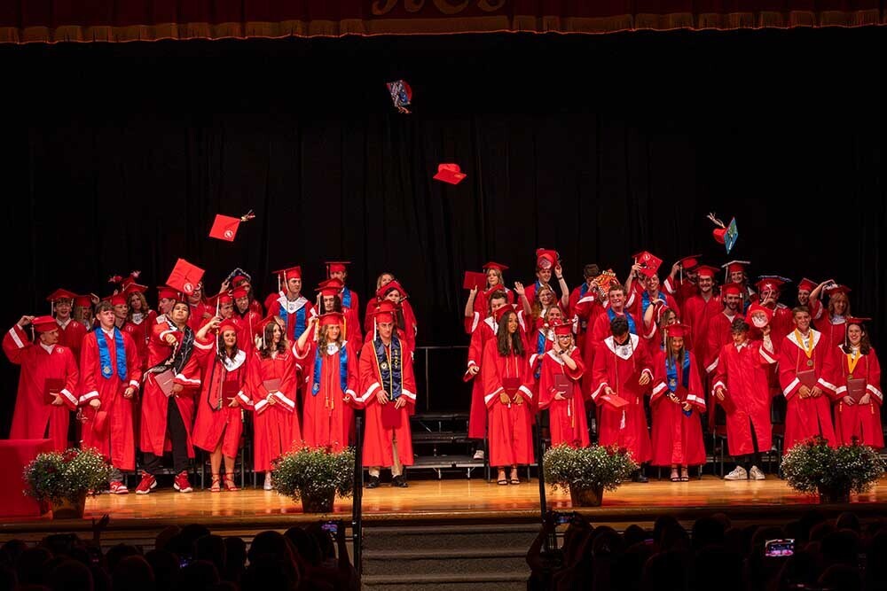 Dozens of graduates in red gowns stand on stage as some throw their caps into the air