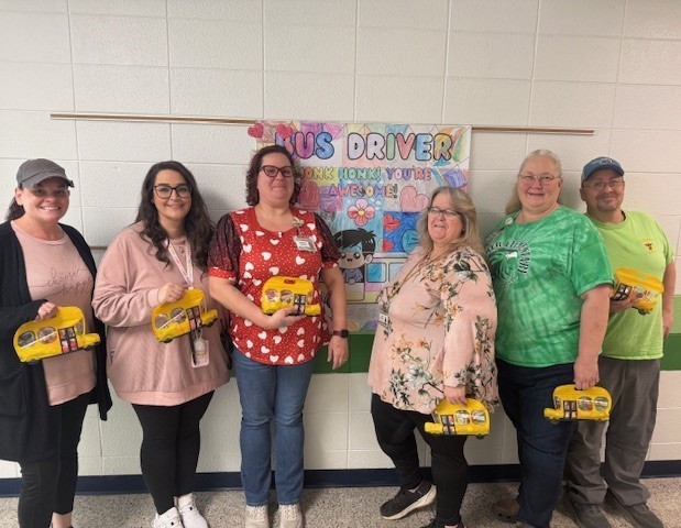 Ms. Branson, Ms. Johnson, Ms. Petersen, Ms. Marshall, Ms. Mitchell and Mr. Davidson standing in front of a bus driver poster.