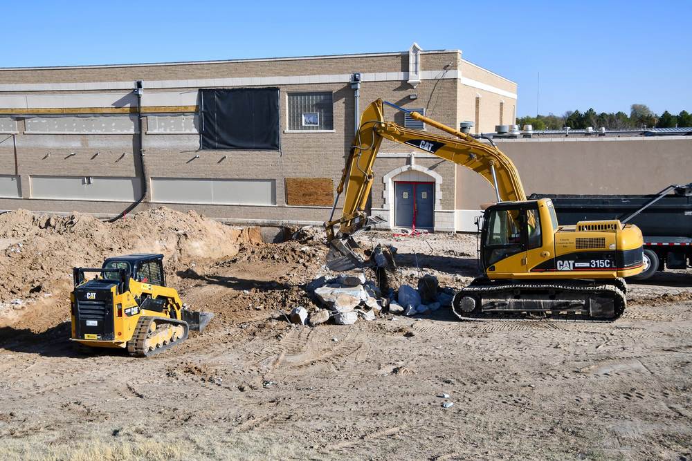 Excavator and compact track loader at a construction site outside a brick school building, with dirt piles and broken concrete in the foreground.