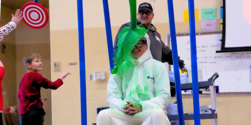 A man in a white suite sits in front of a blue device while getting green slime dumped on his head.